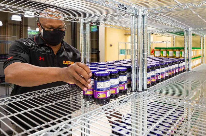 A person wearing a mask and a Maryland shirt stocks jars of food onto chrome pantry shelves.