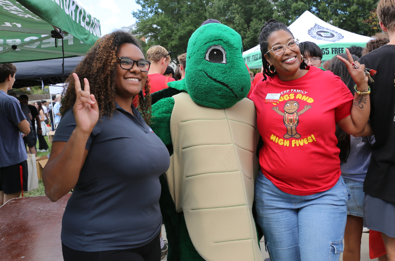Two women and a turtle mascot pose for a photo, giving peace signs at an outdoor event.