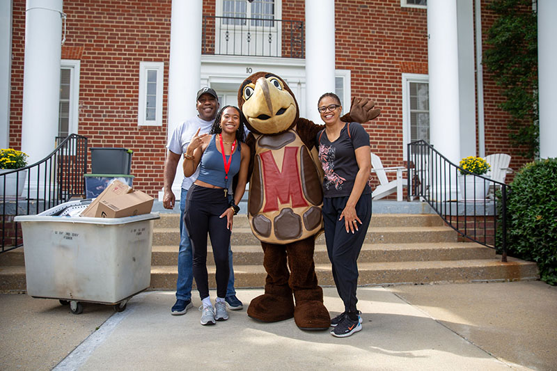 Testudo mascot stands with a smiling student and her parents in front of a house during move-in day.