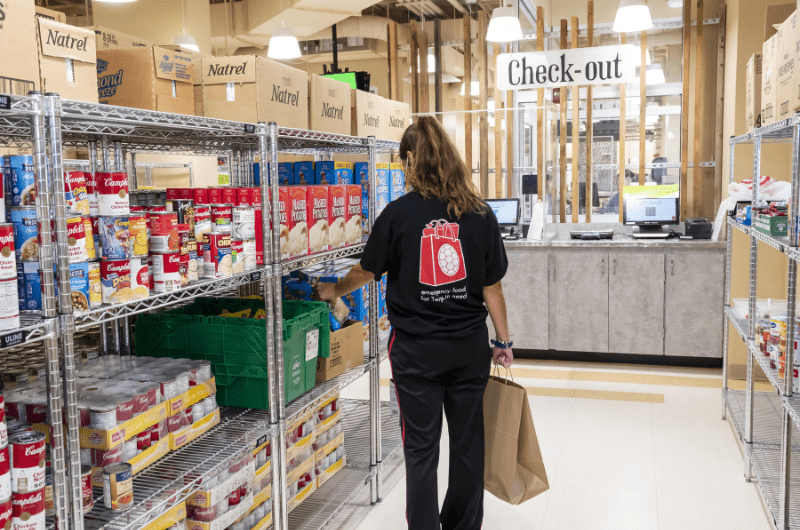 A person shops for groceries in a food pantry, with shelves stocked with canned and boxed goods. A "Check-Out" sign is visible in the background.