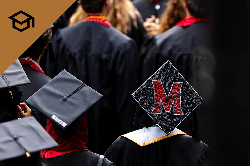 A black graduation cap decorated with a large red and white "M" in a group of graduates.