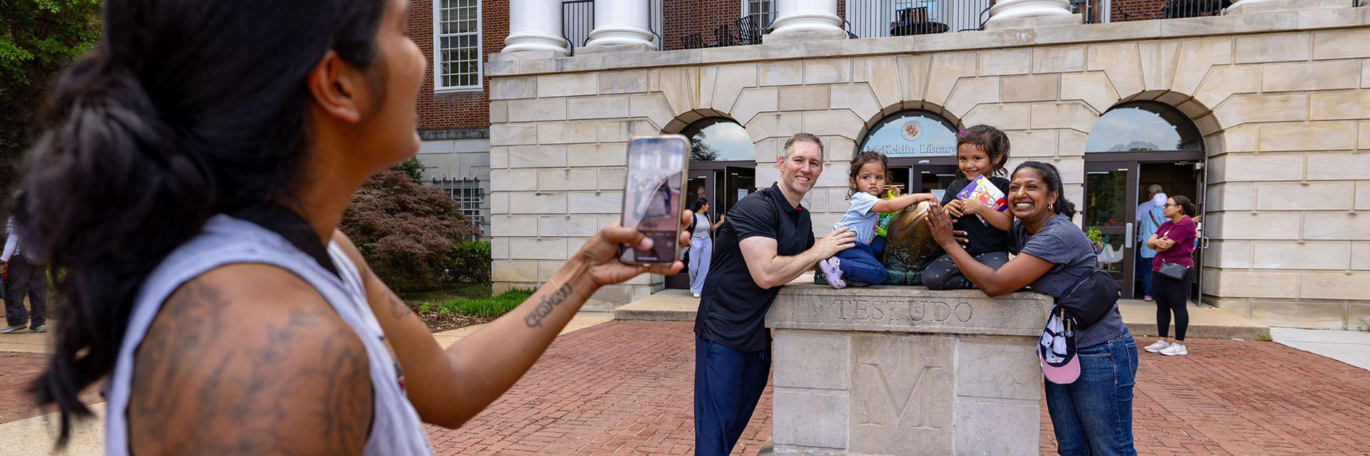  A person takes a phone photo of a smiling family posing with the Testudo statue base outside McKeldin Library.