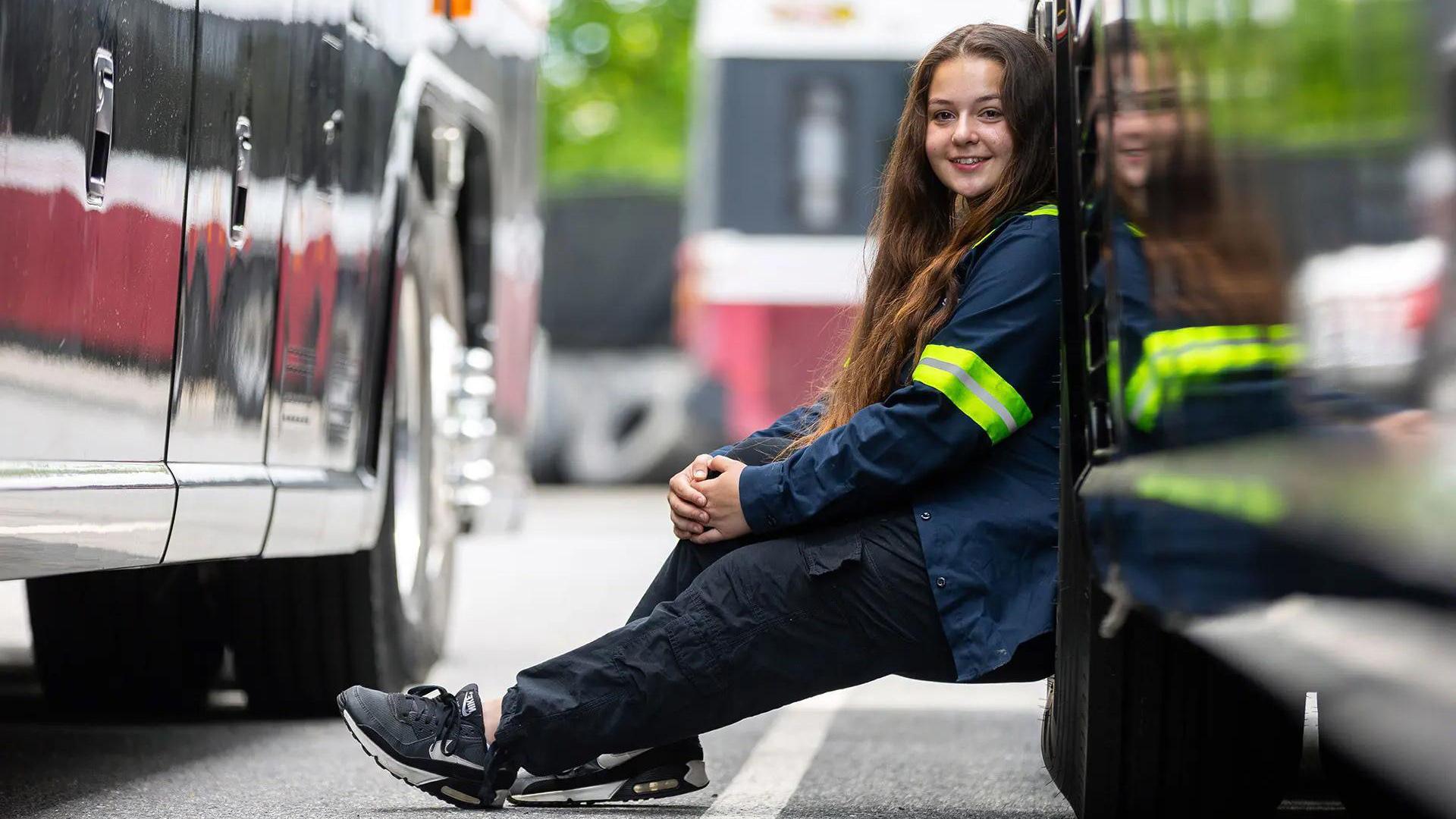 A young woman with long brown hair, wearing a work uniform with a reflective jacket, sits leaning against a bus tire. 