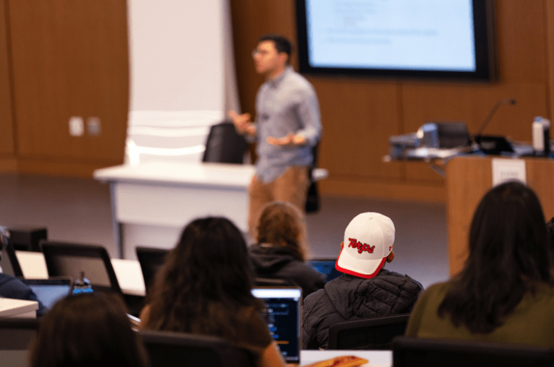  A blurry instructor lectures a class while a student in the foreground wears a white cap with red lettering.