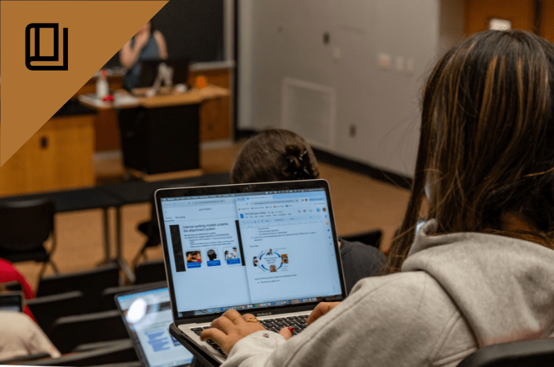 A student works on a laptop in a classroom, with the screen displaying a presentation.