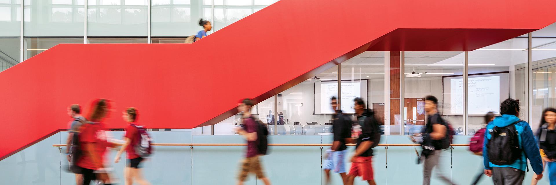 Students walking through Edward St. John building with a bold red staircase and glass walls.