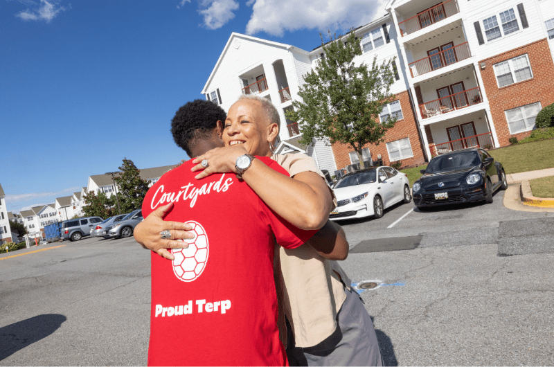 Two people embrace in an outdoor apartment complex parking lot on a sunny day.
