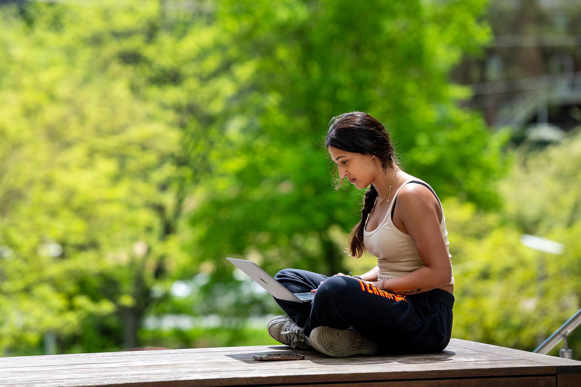 Student sitting outdoors on campus, using a laptop on a sunny day surrounded by green trees.