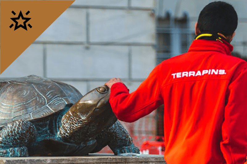  A person in a red jacket with "TERRAPINS" on the back touches the nose of a bronze Testudo statue.