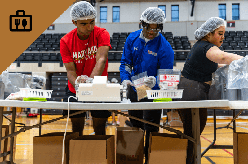 Three people wearing hairnets and gloves pack food into bags on a table.