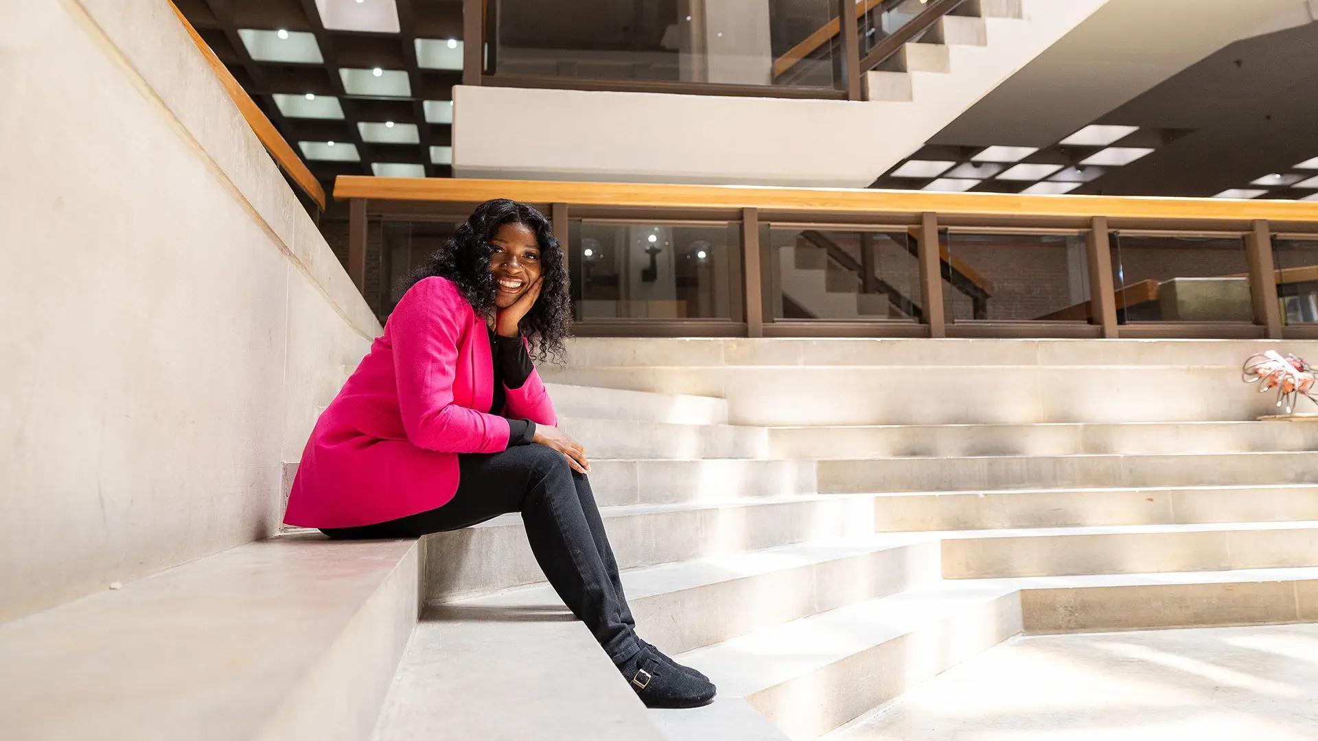 Student wearing a bright pink jacket, smiling while sitting on indoor concrete steps in a campus building.