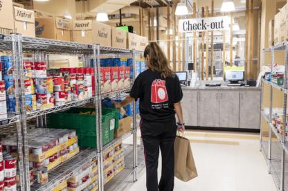 A person shops for groceries in a food pantry, with shelves stocked with canned and boxed goods. A "Check-Out" sign is visible in the background.